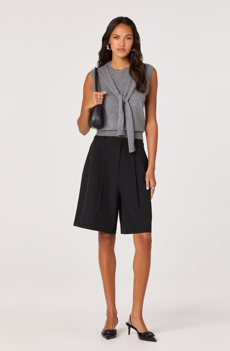 A woman is wearing a sleeveless gray top with a tie front detail, paired with high-waisted, loose-fitting black shorts that reach just above the knees. She accessorizes with hoop earrings, a black shoulder bag, and black pointed-toe heels with a bow detail.