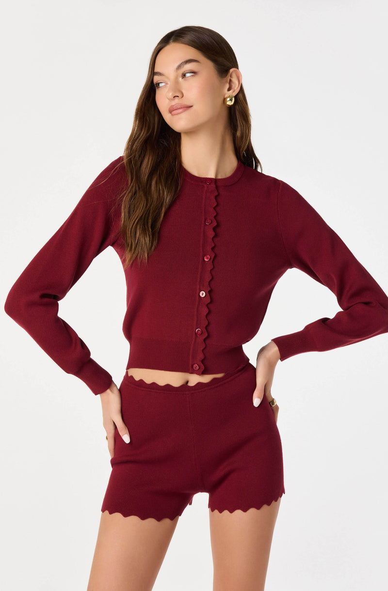A woman is wearing a matching maroon outfit consisting of a long-sleeved, button-up cardigan with a wavy trim along the front and cuffs, and a pair of high-waisted shorts with a scalloped hem. The cardigan is slightly cropped, revealing a small section of her midriff. She has long, wavy brown hair and is accessorized with gold earrings and a ring.