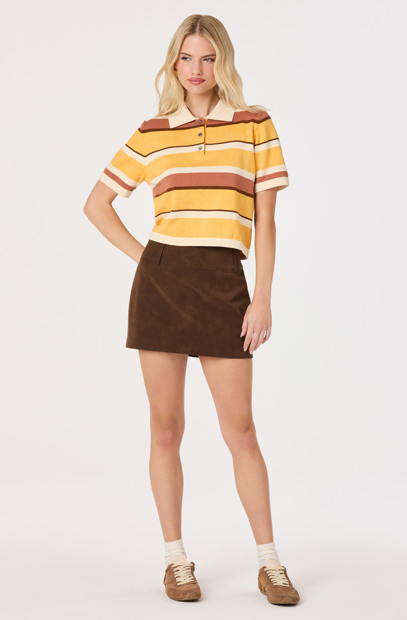 Woman wearing a striped yellow, brown, and cream short-sleeve polo shirt with a collar, and a brown skirt, paired with beige sneakers and white ankle socks.