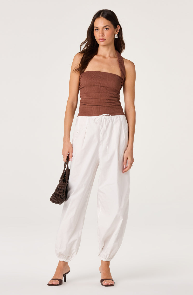 Woman in brown strap top, white baggy pants, and black heels, holding a dark brown purse.