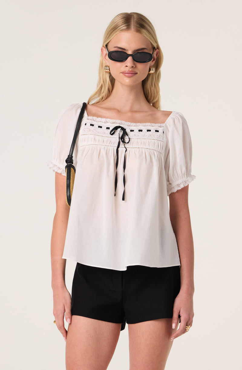 Woman in white blouse with black lace-up detail, black shorts, and oval sunglasses.