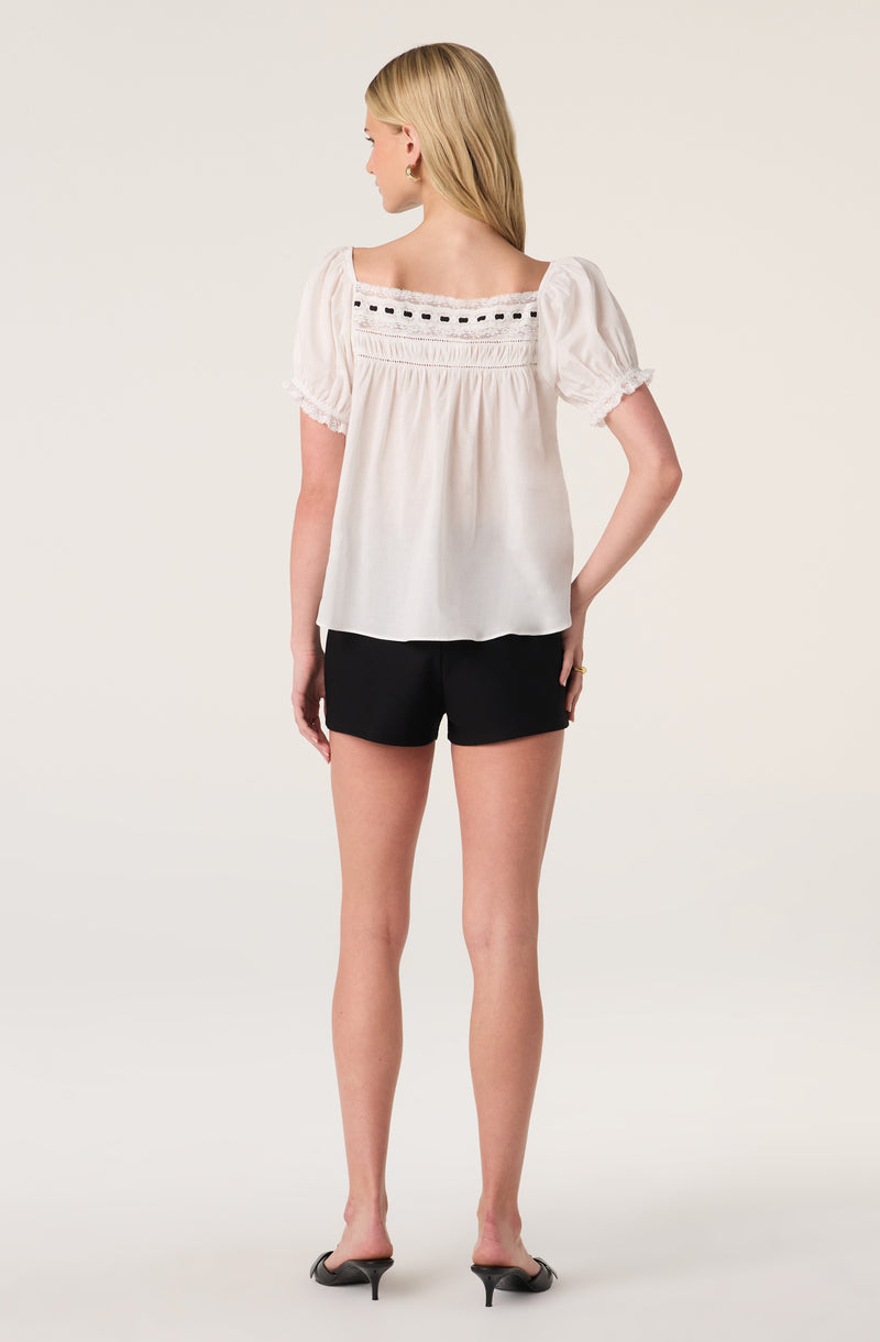 Woman wearing a white blouse with puffed sleeves and black shorts, standing with her back facing the camera.