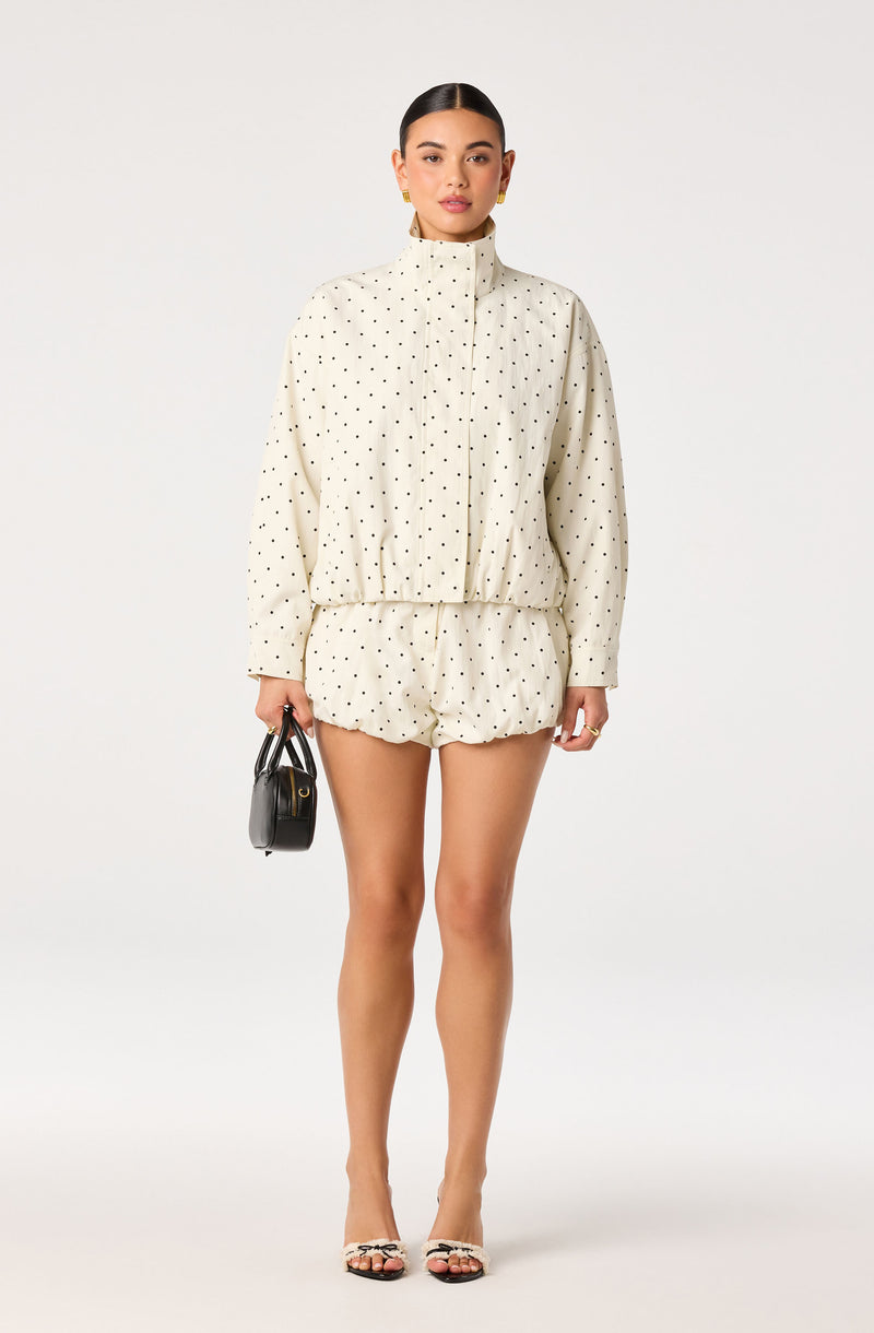 Woman wearing a cream polka dot hoodie and shorts, holding a small black handbag.
