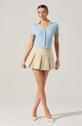 A woman is wearing a light blue short-sleeved button-up sweater with a textured, open-knit pattern and a collar. She pairs it with a beige, high-waisted, pleated mini skirt. She completes the look with beige pointed-toe heels.