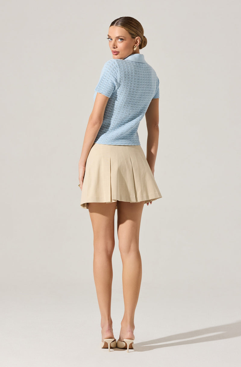 A woman is wearing a light blue, short-sleeved, textured knit top with a collar, paired with a beige pleated mini skirt and beige high-heeled shoes.