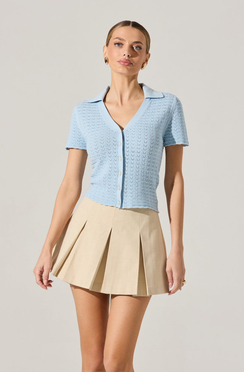 A woman is wearing a light blue, short-sleeved, button-up knit top with a collar. She pairs it with a beige pleated skirt.