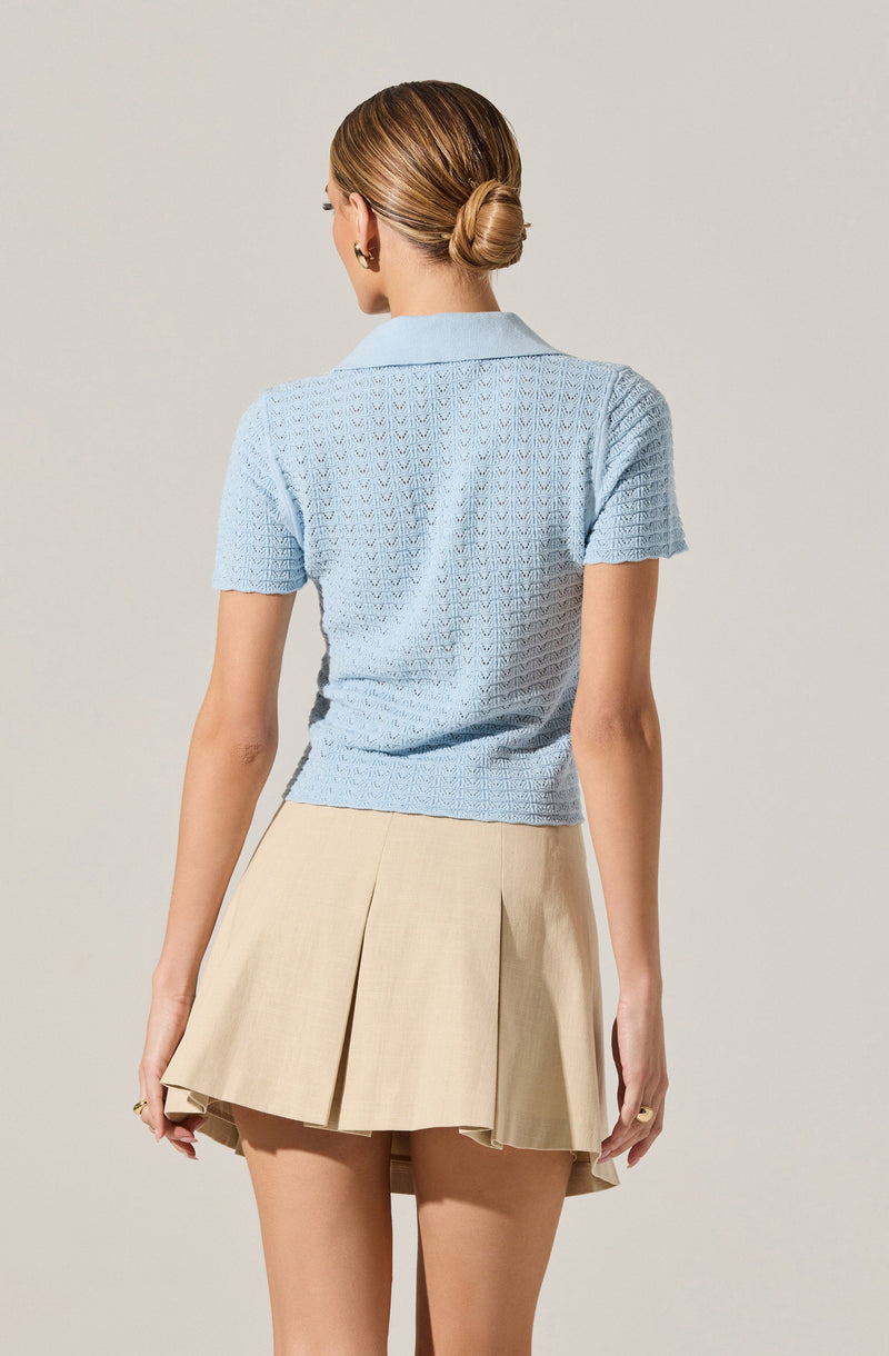 A woman is wearing a light blue textured short-sleeve knit top with a collar, paired with a beige pleated skirt.