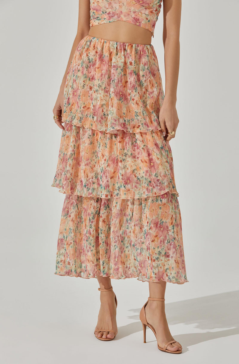 A woman is wearing a flowy, tiered maxi skirt with an abstract floral pattern in shades of pink, orange, yellow, and green. She pairs it with nude high-heeled sandals and a matching crop top made from the same fabric.