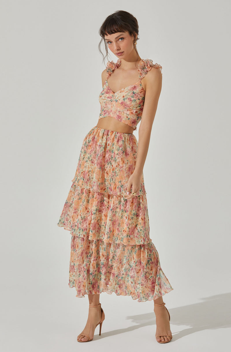 A woman is wearing a two-piece floral outfit with a sleeveless crop top and a high-waisted, tiered maxi skirt. The top has ruffled shoulder straps and a V-neckline, featuring a pink and peach floral pattern. The skirt has the same floral pattern, with multiple ruffled tiers that create a flowing silhouette. She is also wearing strappy nude high-heeled sandals.