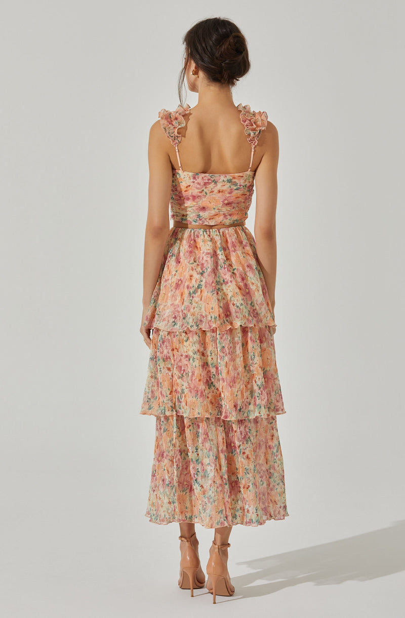 A woman is wearing a sleeveless, floral-print dress with ruffled shoulder straps and a fitted bodice. The dress has a tiered, flowing skirt that reaches ankles, featuring a multicolored pattern with shades of pink, peach, green, and cream. She is also wearing beige high-heeled shoes with ankle straps.