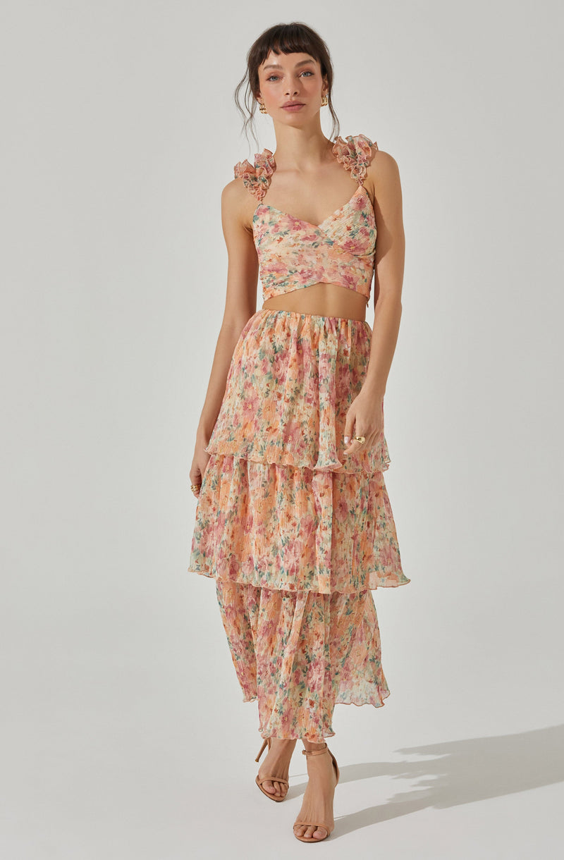 A woman is wearing a sleeveless, floral-print crop top with ruffled shoulder details and a matching high-waisted, tiered skirt that reaches her ankles. The fabric has a soft, watercolor-like floral pattern in shades of pink, peach, and green.