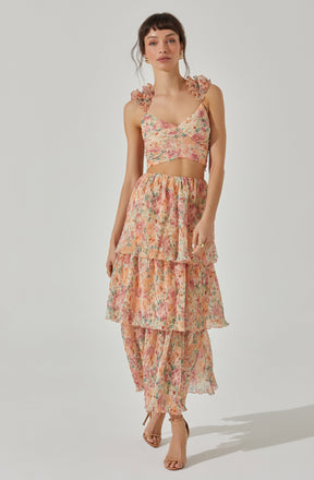 A woman is wearing a sleeveless, floral-print crop top with ruffled shoulder details and a matching high-waisted, tiered skirt that reaches her ankles. The fabric has a soft, watercolor-like floral pattern in shades of pink, peach, and green.