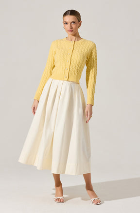 A woman is wearing a yellow, long-sleeve, button-up cardigan with a cable knit pattern. She pairs it with a high-waisted, long, cream-colored skirt that has pleats and a fluttery, flowing silhouette. She is also wearing white open-toe sandals with a small heel.
