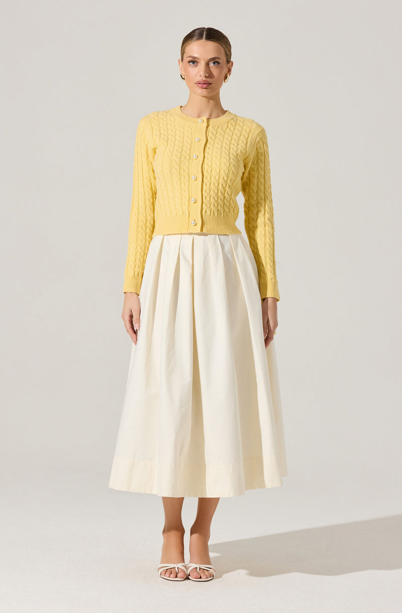 A woman is wearing a light yellow, long-sleeved, cable-knit cardigan with a round neckline and button closure down the front. She pairs it with a high-waisted, cream-colored, wide-legged midi skirt with pleats. She completes the outfit with open-toe, strappy heels.