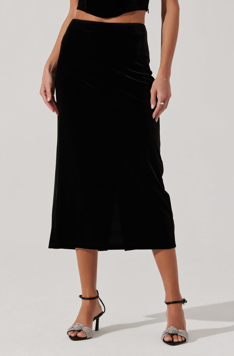 A woman is wearing a black velvet cropped top and a high-waisted, ankle-length black velvet skirt. She is also wearing black ankle-strap high heels with decorative bows on the toes.
