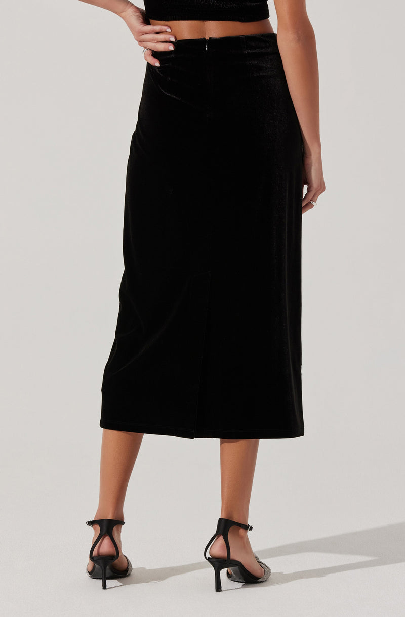 The image shows a person wearing a black, high-waisted, ankle-length skirt made of velvet material. The skirt features a slight slit at the back. They are also wearing black high-heeled sandals with thin straps. The person's top is a black cropped garment that exposes their midriff.