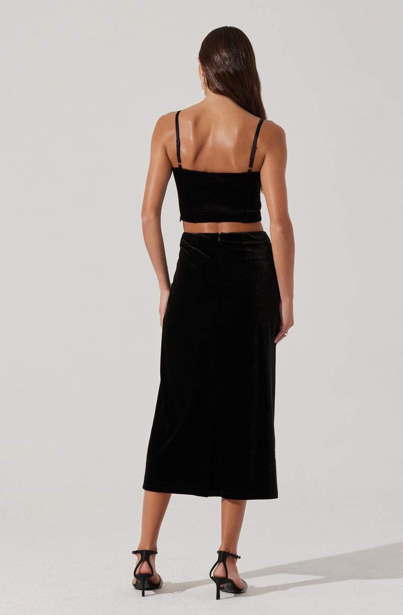 A woman is wearing a black spaghetti strap crop top with a textured fabric, paired with a high-waisted, full-length black skirt made of velvet. She is also wearing black strappy high-heeled sandals.