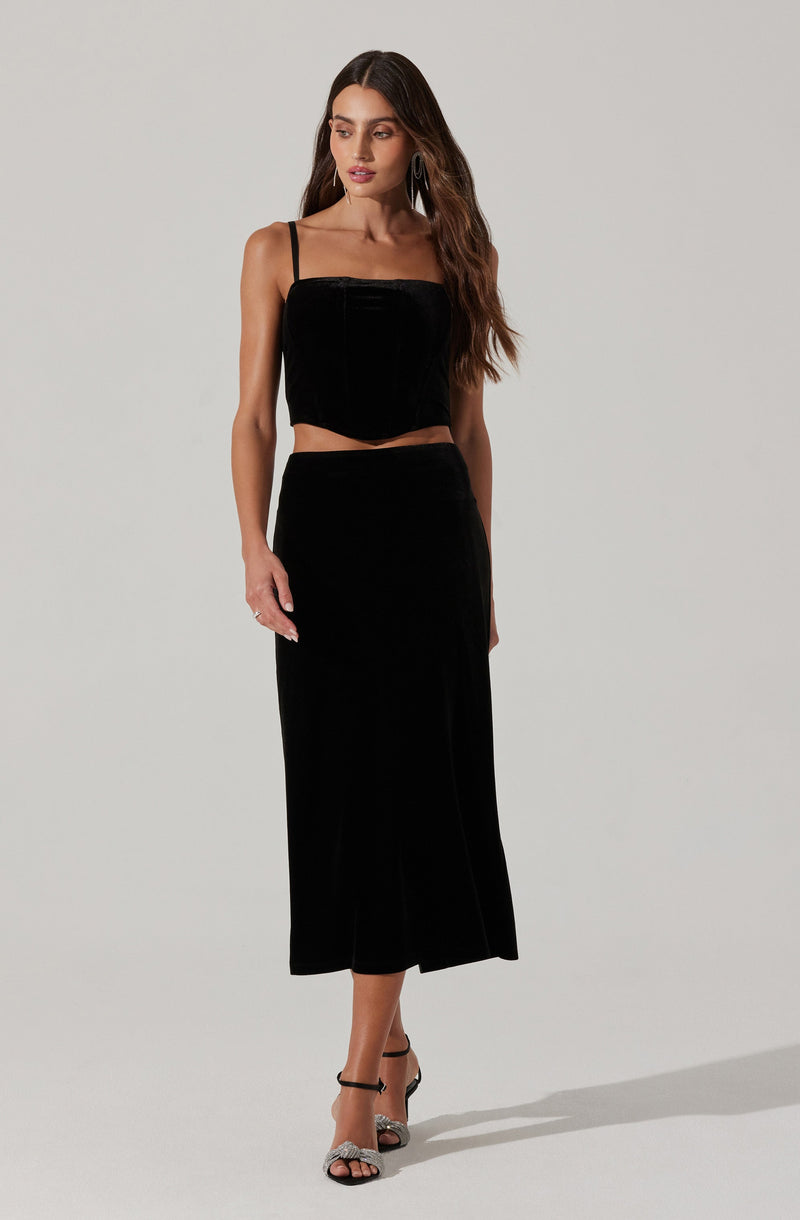 A woman is wearing a black sleeveless cropped top with thin spaghetti straps and a high-waisted, ankle-length black skirt. The top has a square neckline and appears to be made of a velvety fabric. The skirt is fitted at the waist and has a subtle vertical stripe pattern, with a slightly flared hem. She is also wearing open-toe black and silver high heels with ankle straps.