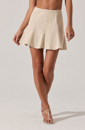 A woman is wearing a high-waisted, cream-colored A-line mini skirt made of a smooth, slightly shiny fabric with vertical seam details. She is also wearing white strappy high-heeled sandals.