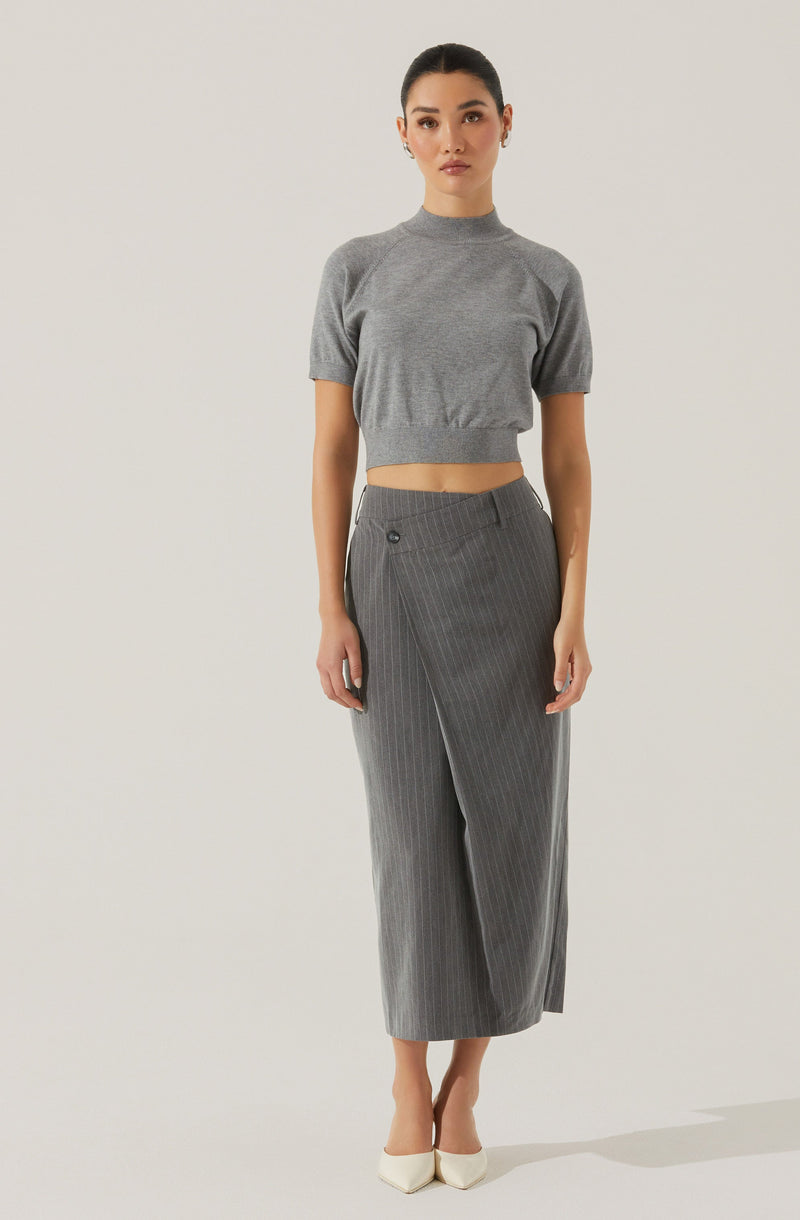 A woman is wearing a short-sleeved, gray knitted crop top with a high neckline, paired with high-waisted, gray wide-leg trousers featuring a subtle pinstripe pattern and front pleats. She is also wearing light-colored pointed-toe heels.