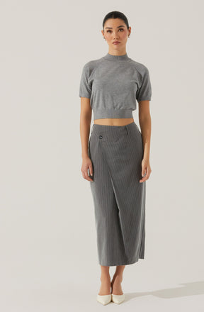 A woman is wearing a short-sleeved, gray knitted crop top with a high neckline, paired with high-waisted, gray wide-leg trousers featuring a subtle pinstripe pattern and front pleats. She is also wearing light-colored pointed-toe heels.