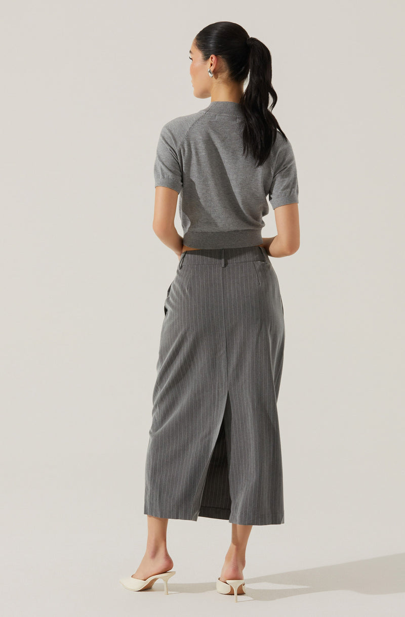 A woman is wearing a short-sleeved, light grey knit top paired with high-waisted, wide-leg, grey pinstripe trousers that have a back slit. She is also wearing white, pointed-toe high heels with a small heel. Her dark hair is styled in a high ponytail.
