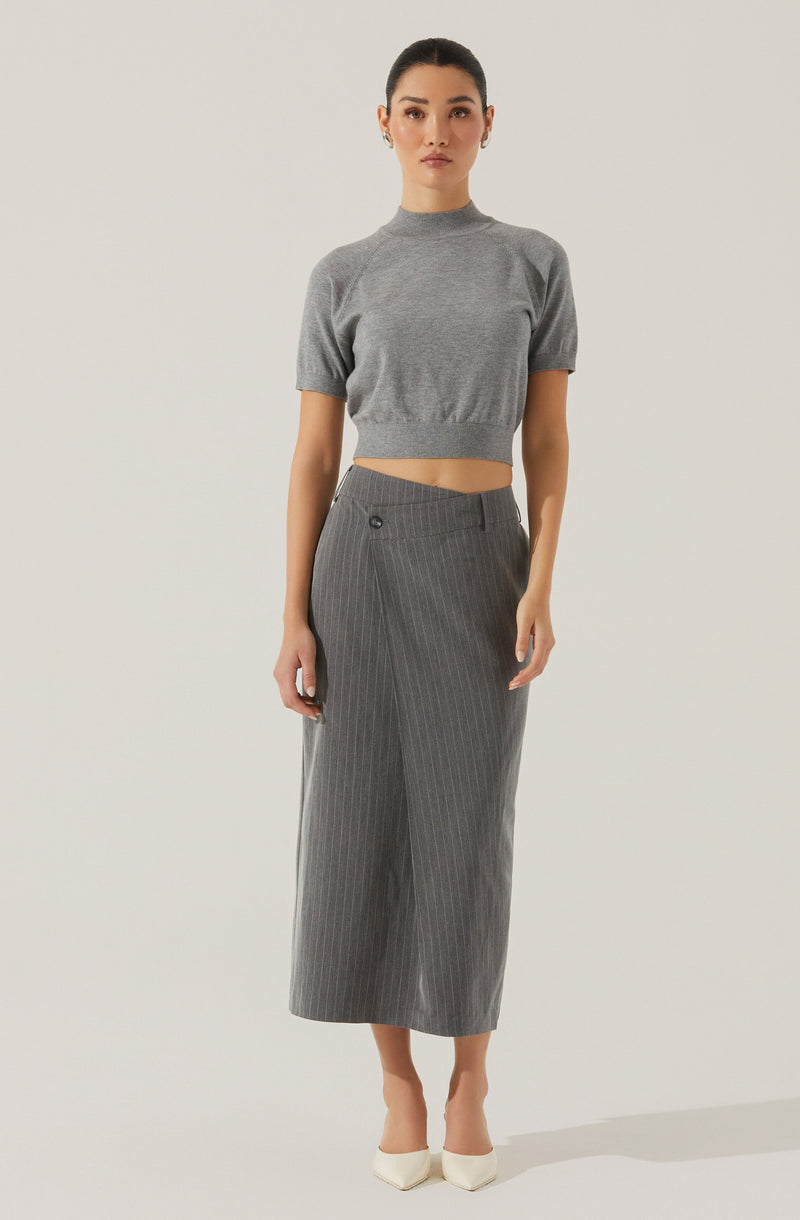 A woman is wearing a grey, short-sleeved, cropped sweater with a high neckline, and high-waisted, wide-leg grey pinstripe trousers that reach her mid-calf. She is also wearing white pointed-toe shoes.