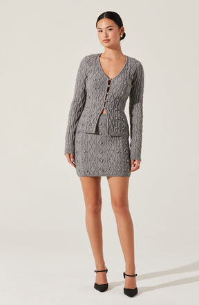 The woman is wearing a coordinated gray knit outfit consisting of a cable-knit cardigan with a V-neckline and button closure, and a matching mini skirt with a diamond and cable-knit pattern. She is also wearing black ankle-strap high heels.