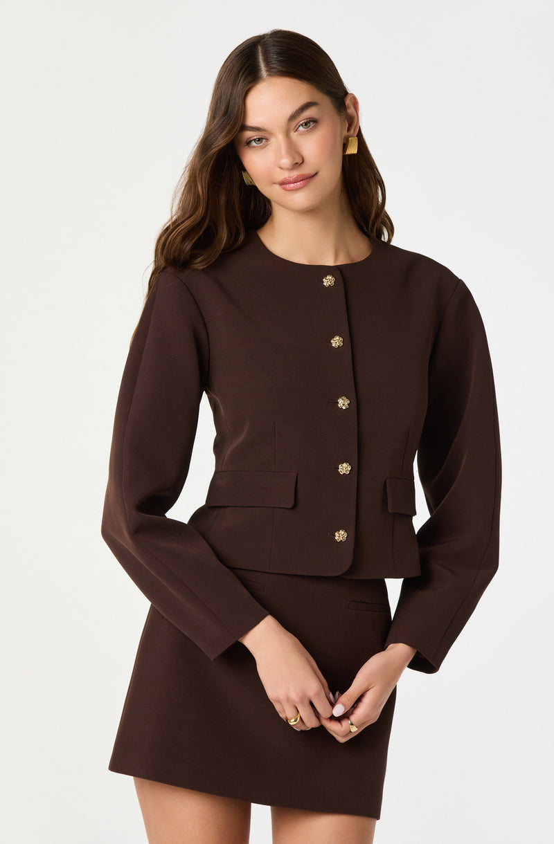A woman is wearing a dark brown, fitted, long-sleeve blazer with a round neckline and gold decorative buttons down the front. The blazer has a slightly cropped hem and features small flap pockets on each side. She is also wearing a matching dark brown skirt that is short and straight.