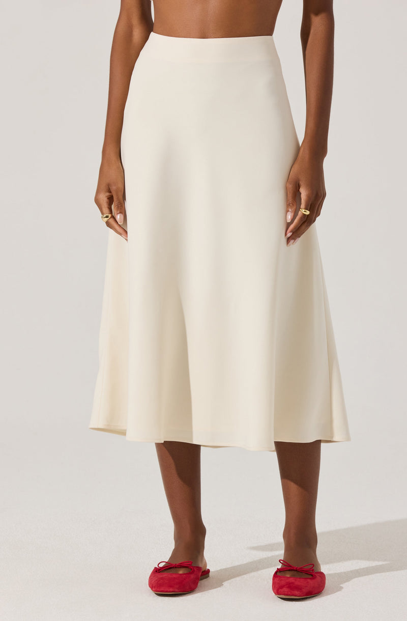 A woman is wearing a high-waisted, ankle-length, cream-colored skirt made of a smooth, flowy fabric. She is also wearing bright red ballet flats with bows on the front. Her hands are resting at her sides and she has some rings on her fingers.