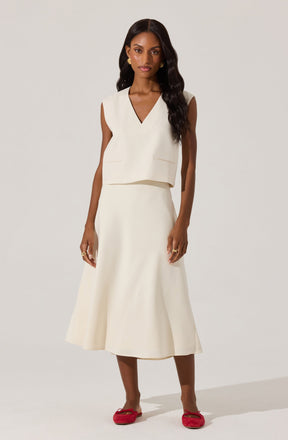 A woman is wearing a sleeveless, off-white top with a V-neckline and two small slit pockets on the front. She pairs it with a matching off-white, A-line midi skirt. She completes her outfit with red ballet flats that have a bow detail on the front.
