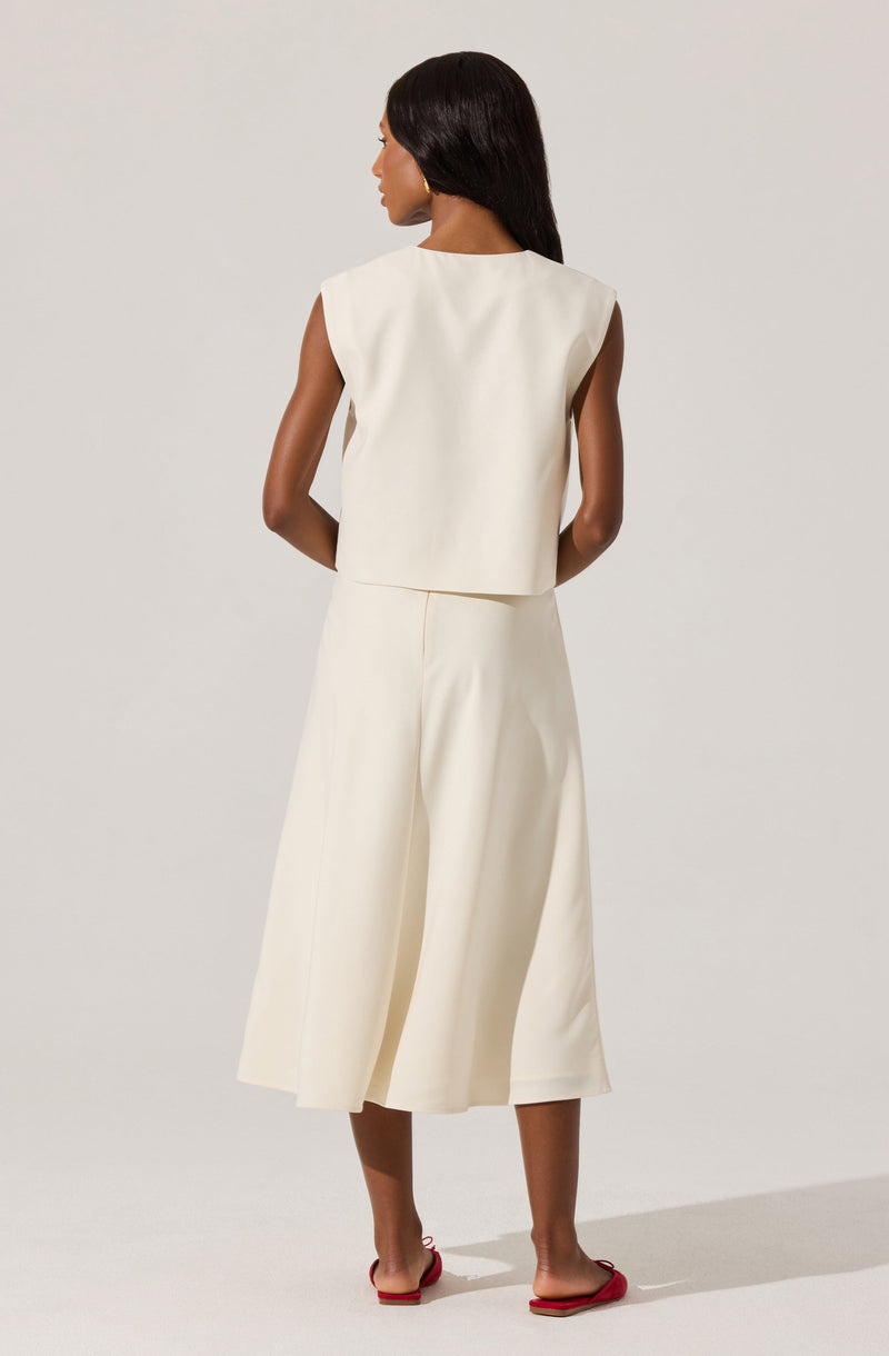 A woman is wearing a sleeveless, cream-colored top with broad shoulder panels and matching wide-leg, cropped pants made from a smooth, structured fabric. She is also wearing red flat sandals.