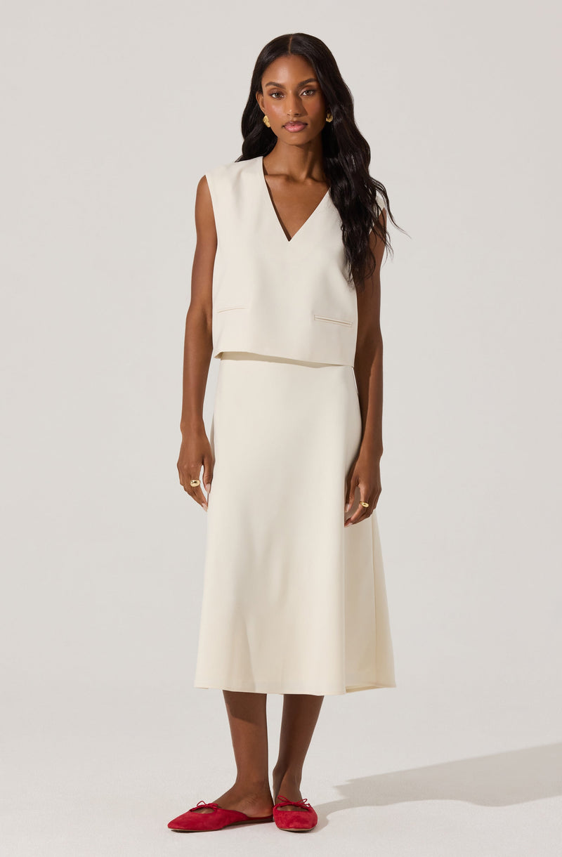 A woman is wearing a sleeveless, cream-colored dress with a V-neckline. The top of the dress is slightly loose and cropped, sitting above the waist of the long, straight skirt. She is also wearing bright red flat shoes with a bow detail.