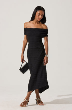 A woman is wearing a black off-the-shoulder, knitted dress that fits snugly to her body and extends down to her ankles. She accessorizes with a silver bracelet, rings, a small black handbag, and strappy high-heeled sandals.
