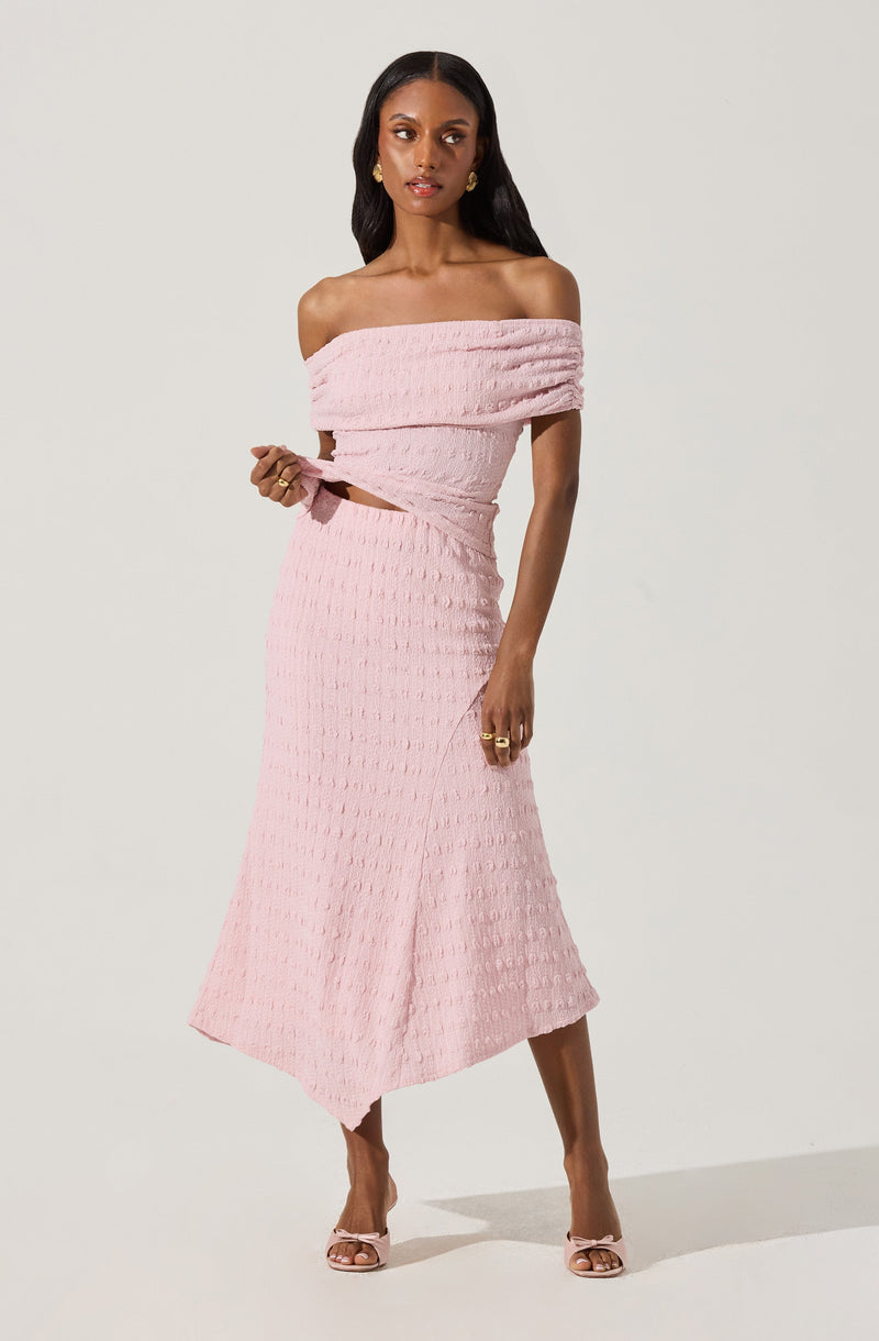 The outfit consists of a light pink, off-the-shoulder dress made from textured fabric. The dress features a fitted bodice with a wide, ruched neckline that wraps around the shoulders. It has a matching pink fabric belt that ties at the waist. The skirt extends to mid-calf length with a slight flare, creating an elegant silhouette. The model is also wearing open-toed, high-heeled shoes in a matching pink shade.