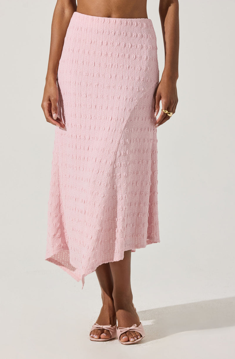A woman is wearing a light pink textured midi skirt with a subtle checkered pattern and an asymmetrical hemline. She has on matching light pink open-toe heels with bows on the front. She is also wearing a gold ring on her left hand.