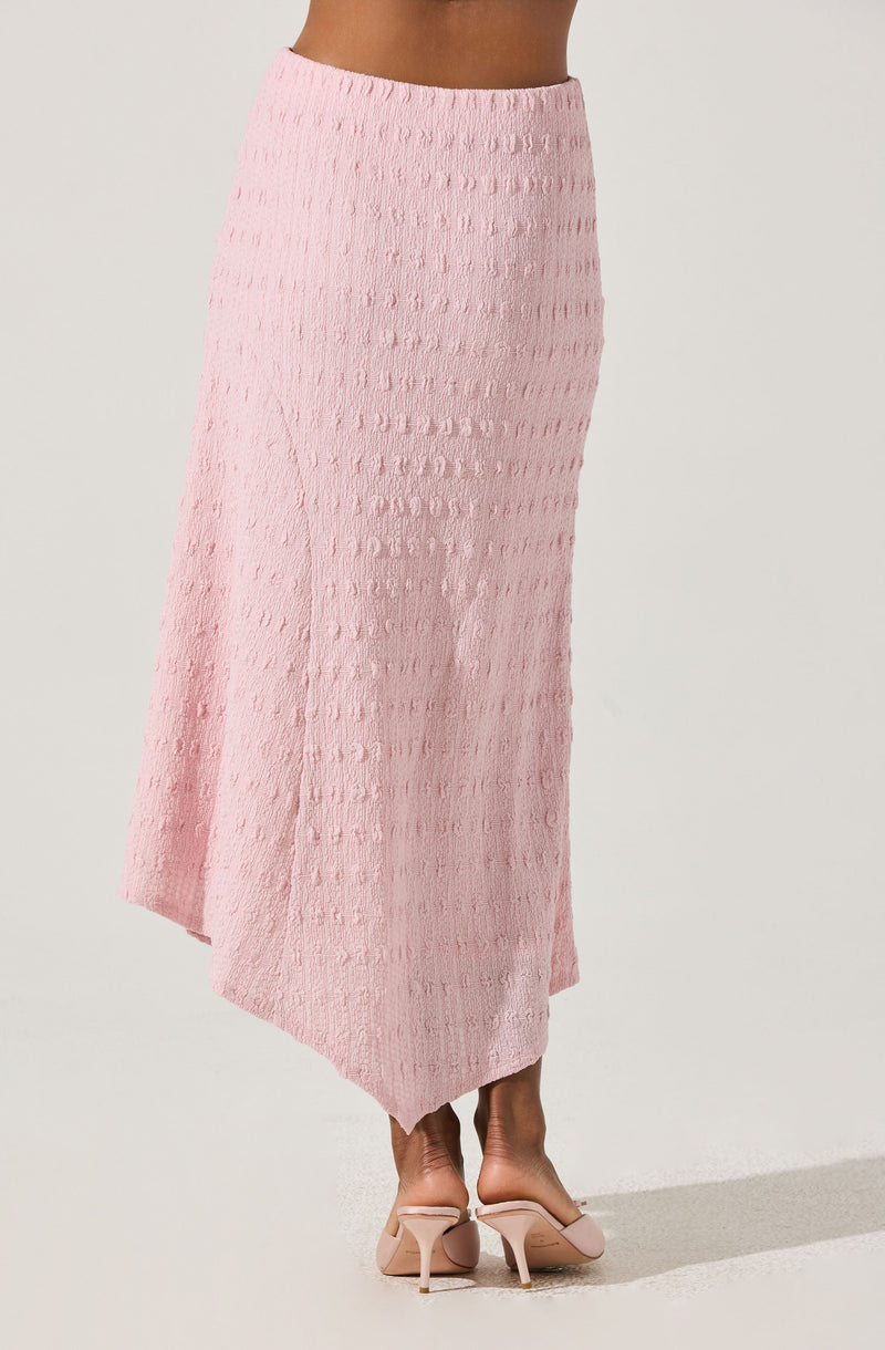 A model is wearing a long, high-waisted pink skirt with a textured, dotted pattern. The skirt has an asymmetrical hemline that is slightly longer at the back. The model is pairing the skirt with nude, open-toe high heels.