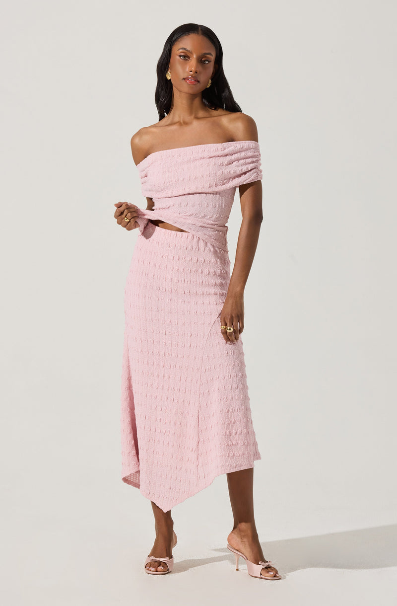 A woman is wearing an off-the-shoulder, light pink textured dress with a matching sash tied at the waist. The dress is midi-length with an asymmetrical hemline that is longer on one side. She is accessorized with gold earrings and rings, and is wearing light pink high-heeled sandals.