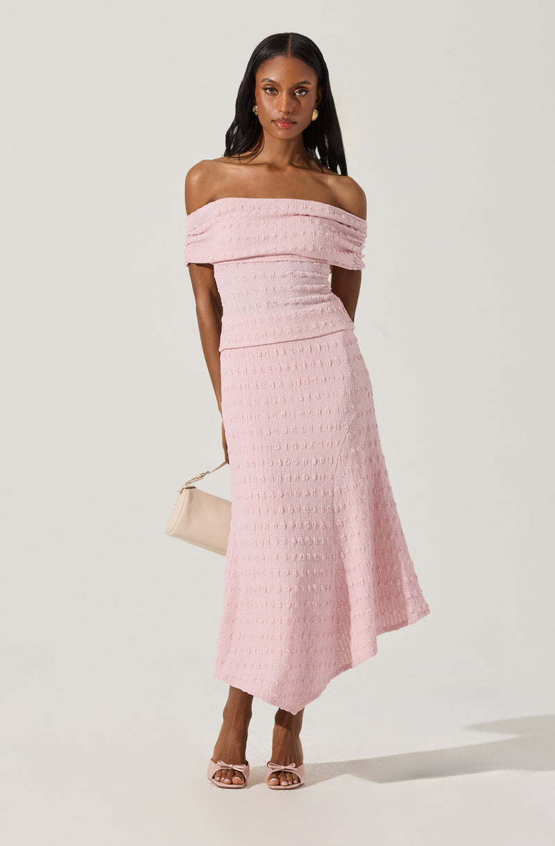 A woman is wearing a light pink off-the-shoulder midi dress with textured fabric and horizontal ruching details. She is holding a beige clutch bag in her left hand and wearing pink open-toe heels with bows.