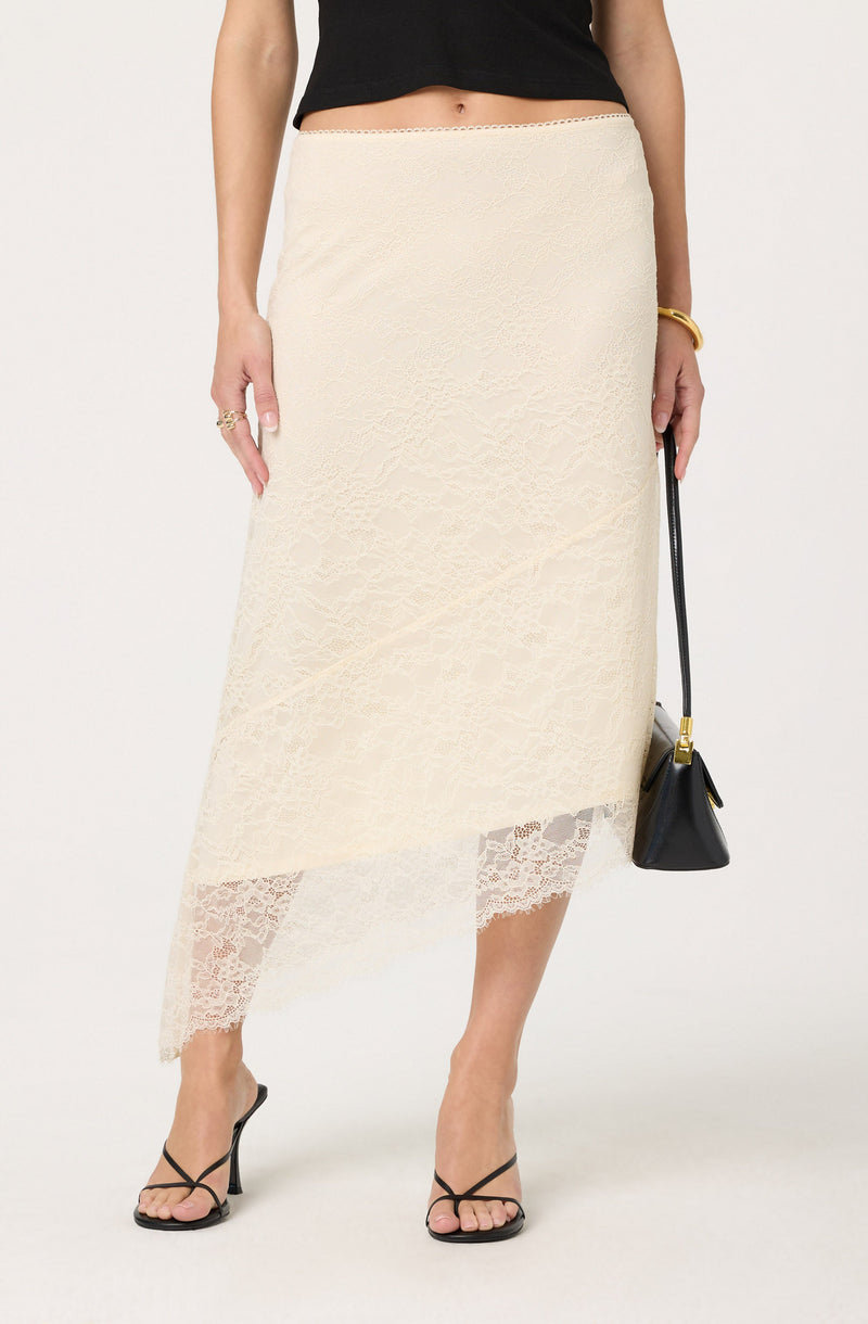 A woman is wearing a black sleeveless top paired with a cream-colored lace skirt that falls below the knees. The skirt features a sheer lace hemline with intricate floral patterns. She accessorizes with a gold bangle, multiple rings, and is holding a black handbag with a gold clasp. She is also wearing black high-heeled sandals with thin straps.