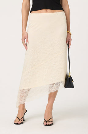 A woman is wearing a black sleeveless top paired with a cream-colored lace skirt that falls below the knees. The skirt features a sheer lace hemline with intricate floral patterns. She accessorizes with a gold bangle, multiple rings, and is holding a black handbag with a gold clasp. She is also wearing black high-heeled sandals with thin straps.