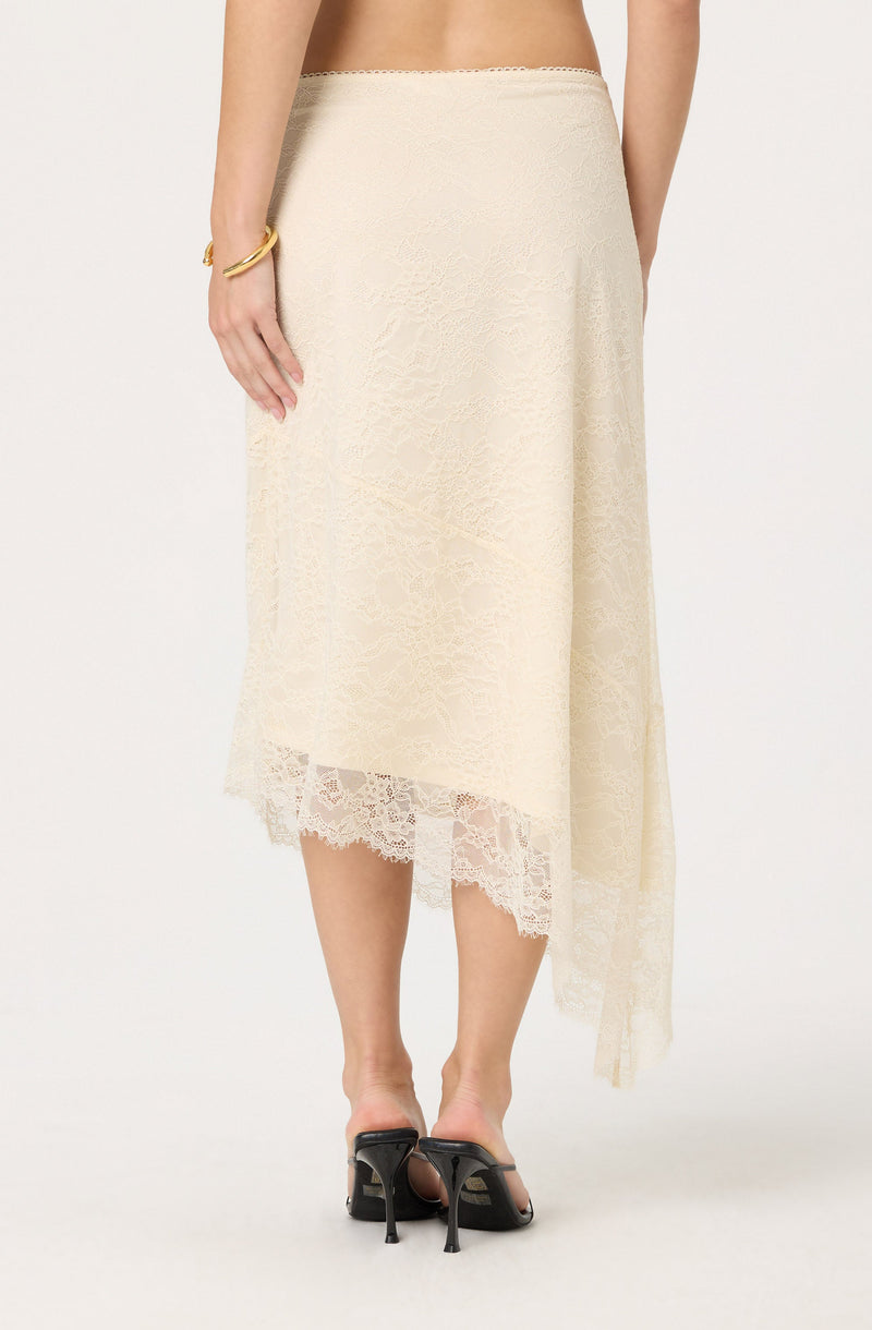 A woman is wearing a cream-colored, asymmetrical lace skirt that falls to mid-calf length. The skirt features a delicate lace overlay with a floral pattern and scalloped lace trim at the hem. She is also wearing black high-heeled sandals with an open toe and ankle strap, and a gold bracelet on her right wrist.