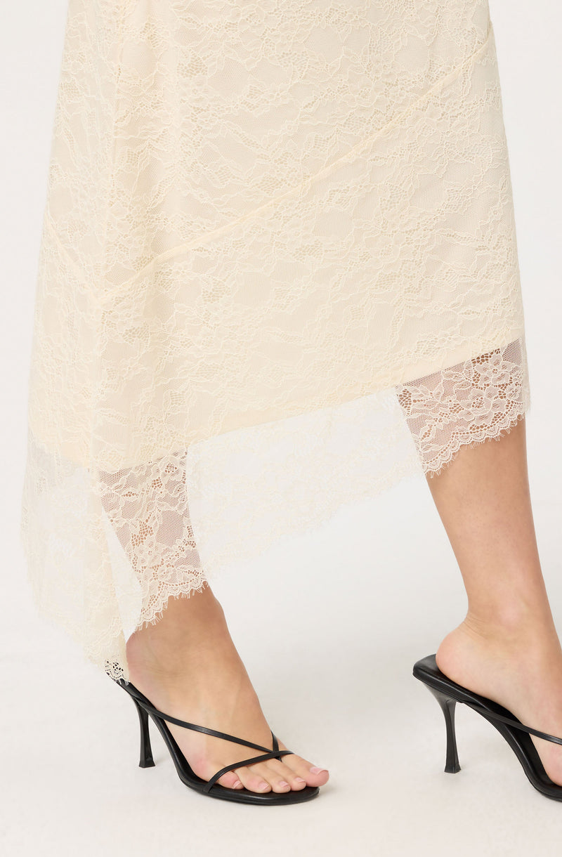 A person is wearing a long, cream-colored lace dress that is slightly sheer, revealing lace-trimmed undergarments beneath. They are also wearing black high-heeled sandals with thin straps over the toes and a stiletto heel.