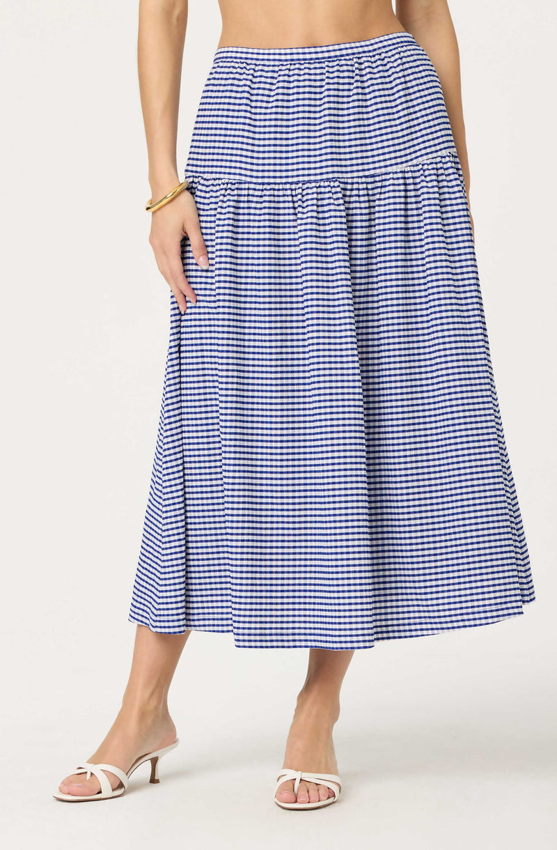 A woman is wearing a blue and white gingham checkered maxi skirt with a high waist. The skirt has a tiered design, with a gathered section at the top and a wider, flowing section below. She is also wearing white high-heeled sandals with a bow detail on the front. She has a gold bracelet on her left wrist.