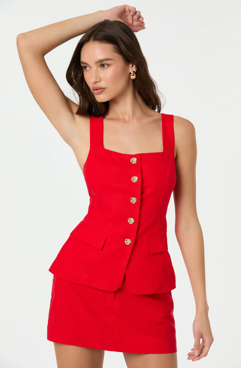 A woman is wearing a sleeveless, fitted red dress with wide shoulder straps. The dress features a square neckline, gold decorative buttons down the front, and two small flap pockets on the front of the skirt.