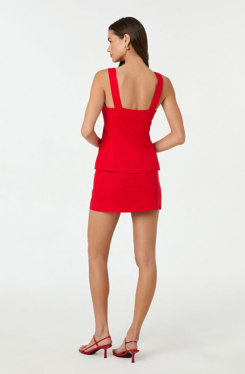 A woman is wearing a sleeveless, bright red two-piece outfit consisting of a fitted top with wide straps and a matching high-waisted mini skirt. She is also wearing open-toed, strappy red high heels.
