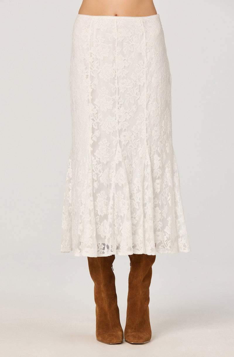 A woman is wearing a long, white lace skirt with a floral pattern that reaches down to her ankles. She pairs the skirt with knee-high brown suede boots. The waistband of the skirt sits at her natural waist.