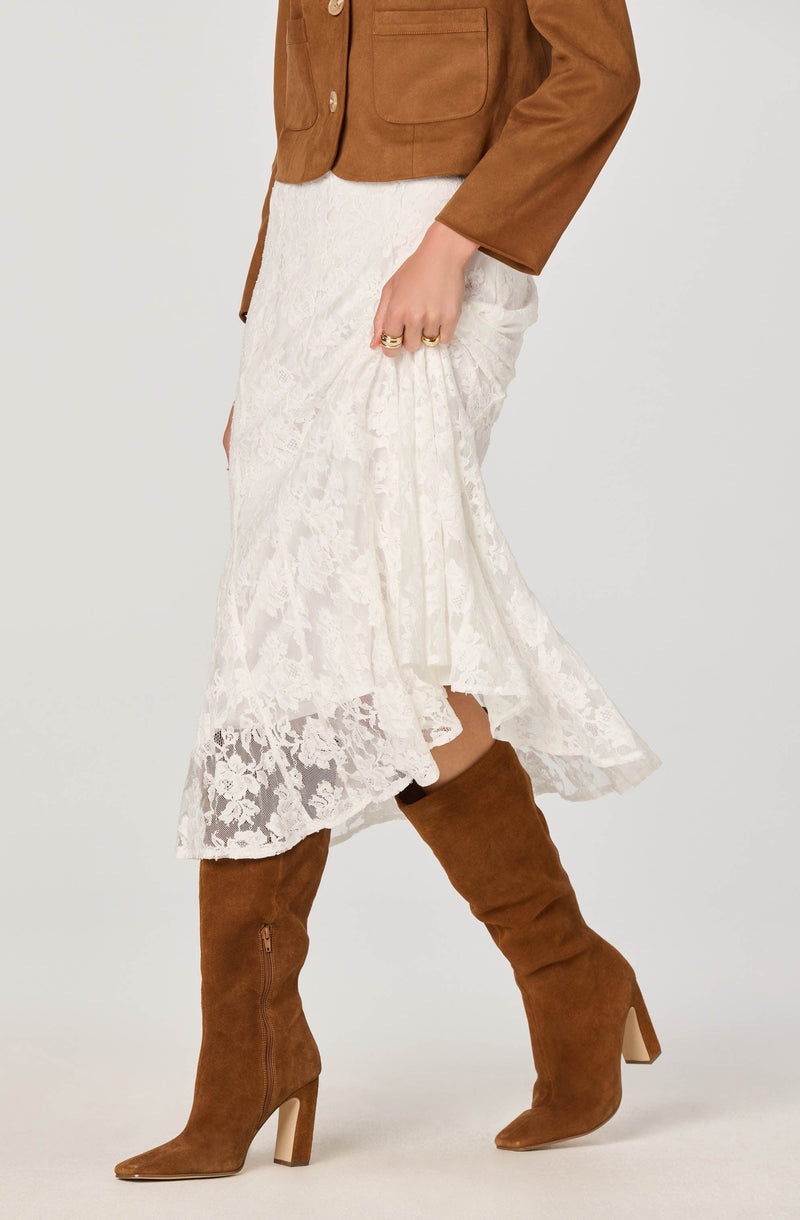 A person is wearing a brown suede jacket with large buttons and patch pockets. They have on a white lace midi skirt with a floral pattern and ruffled hem. They are also wearing knee-high brown suede heeled boots with pointed toes and side zippers. The person has multiple rings on their fingers.