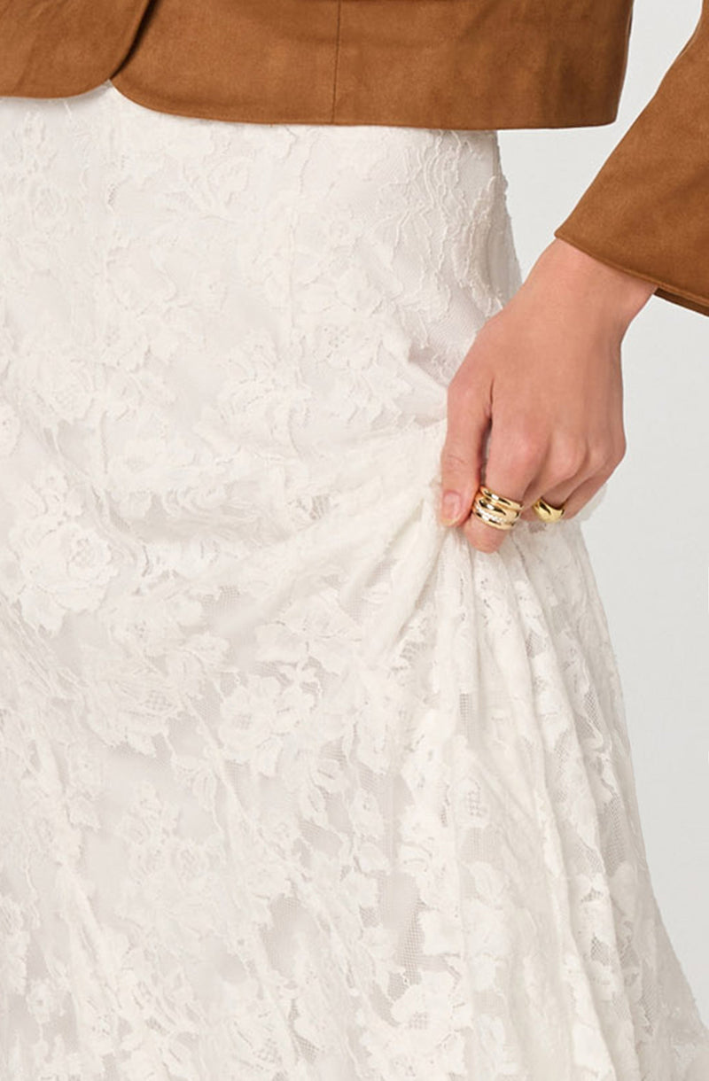 A person wearing a brown jacket and a white lace skirt, with their hand pulling on the skirt. The person is also wearing multiple gold rings on their fingers.