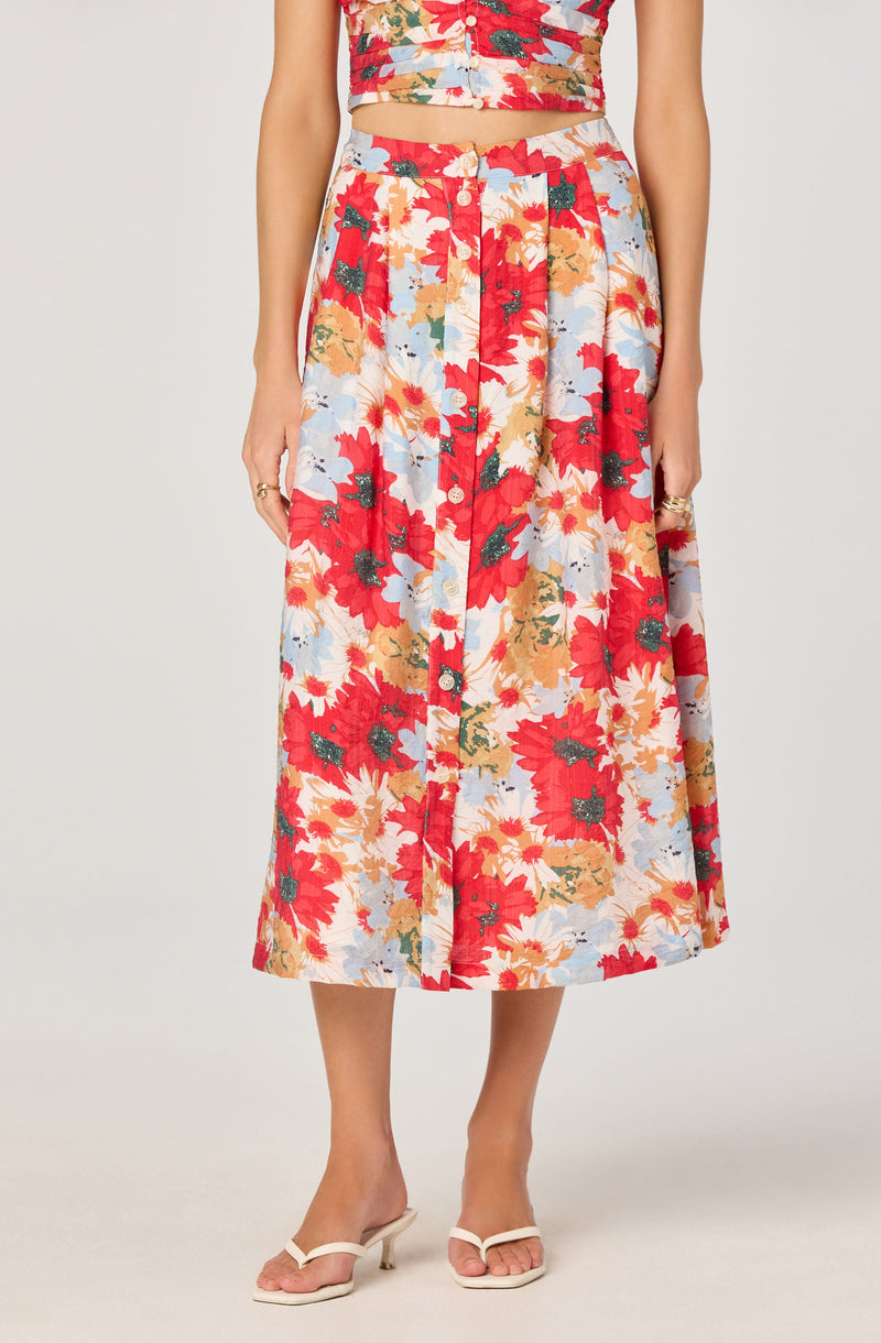 A woman is wearing a matching two-piece outfit consisting of a crop top and a midi skirt. The fabric features a colorful floral pattern with large red, orange, cream, and light blue flowers and green leaves. She is also wearing beige heeled sandals.